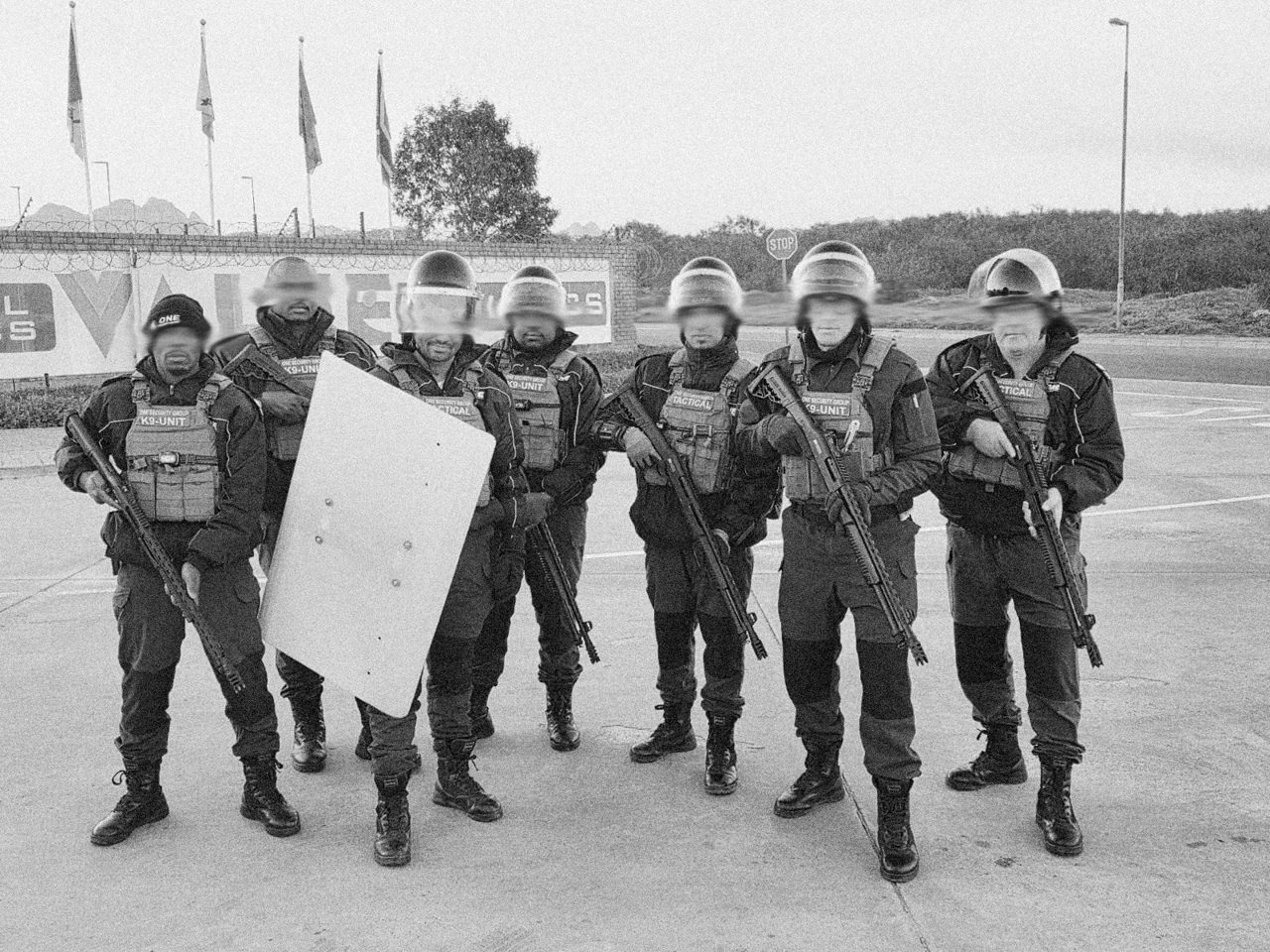 Seven armed ONE tactical and K9 unit officers in helmets and plate carriers, one with a riot shield